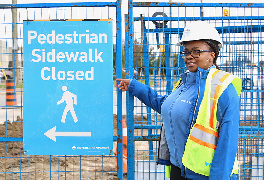 Woman in safety equipment standing in front of a construction fence, pointing to a pedestrian walkway sign